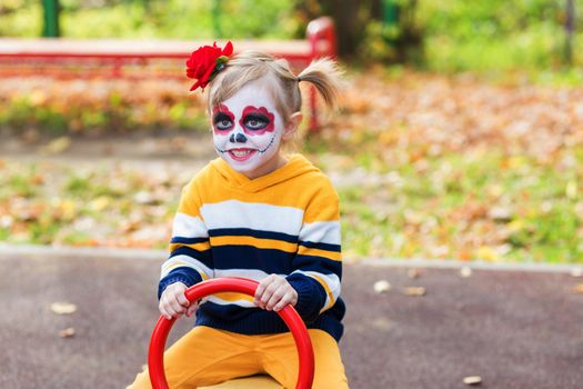 A little preschool girl with Painted Face, smiling at the camera on the playground, celebrates Halloween or Mexican Day of the Dead..