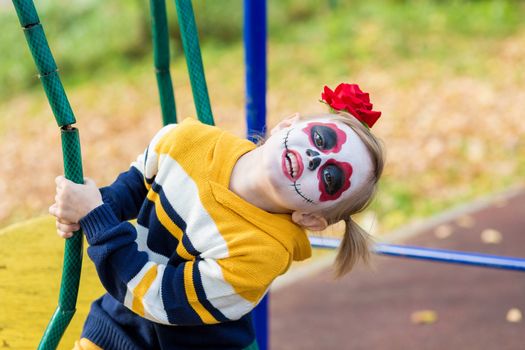 A little preschool girl with Painted Face, shows funny faces on the playground, celebrates Halloween or Mexican Day of the Dead.