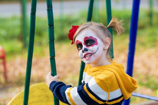 A little preschool girl with Painted Face, shows funny faces on the playground, celebrates Halloween or Mexican Day of the Dead.
