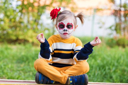 A little preschool girl with Painted Face, sitting on a bench in lotus position on the playground, celebrates Halloween or Mexican Day of the Dead.