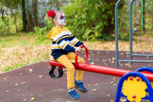 A little preschool girl with Painted Face, rides on a swing in the playground, celebrates Halloween or Mexican Day of the Dead.