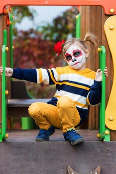 .A little preschool girl with Painted Face, rides a slide on the playground, celebrates Halloween or Mexican Day of the Dead.
