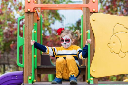 A little preschool girl with Painted Face, rides a slide on the playground, celebrates Halloween or Mexican Day of the Dead.