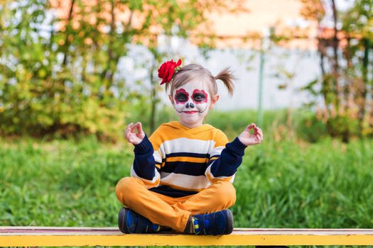 A little preschool girl with Painted Face, sitting on a bench in lotus position on the playground, celebrates Halloween or Mexican Day of the Dead.