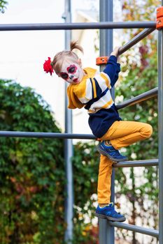 A little preschool girl with Painted Face, climbed the Swedish wall in the playground, celebrates Halloween or Mexican Day of the Dead.