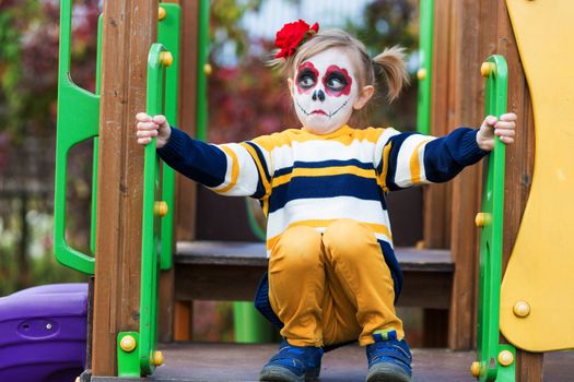 A little preschool girl with Painted Face, shows funny faces on the playground, celebrates Halloween or Mexican Day of the Dead.