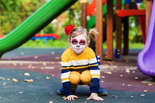 A little preschool girl with Painted Face, smiling at the camera on the playground, celebrates Halloween or Mexican Day of the Dead..