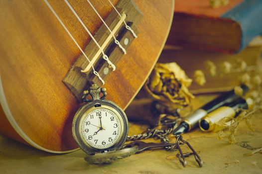 An antique pocket watch leaned against a ukulele and old book with vintage map and brass pen placed on wooden table. closeup and copy space for text. The concept of memories or things in the past.