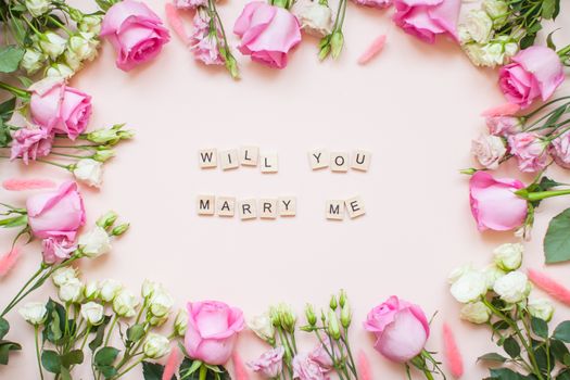 Inscription of wooden blocks will you marry me. Frame of delicate white and pink roses and eustomas on a light pink background. Layout. Flat lay