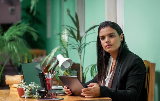 Young woman with long black hair sitting at a working desk with a tablet in her hands.