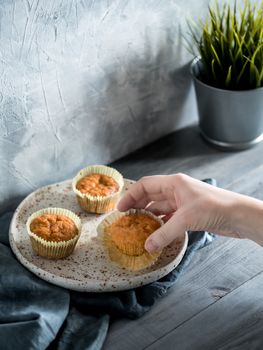 Homemade muffins on craft plate over gray wooden table. Hand hold carrot cupcake. Copy space. Toned image in scandinavian style.