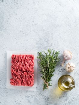 Fresh raw minced beef, fresh rosemary, garlic, olive oil on backing paper over light gray cement background with copy space. Top view or flat-lay.