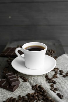 Coffee cup with cookies and chocolate with scattered coffee beans on linen and wooden table background. Mug of black coffee