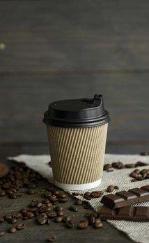 Paper cup of coffee with roasted coffee beans on wooden table. Mug of black coffee with a cookies and chocolate on a table