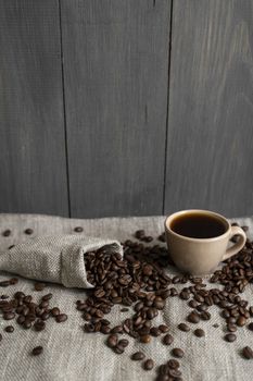Coffee scattered from a linen bag and a cup of black coffee Espresso or Americano standing on a wooden table. Fresh arabica coffee beans on linen textile. Aromatic freshly brewed coffee