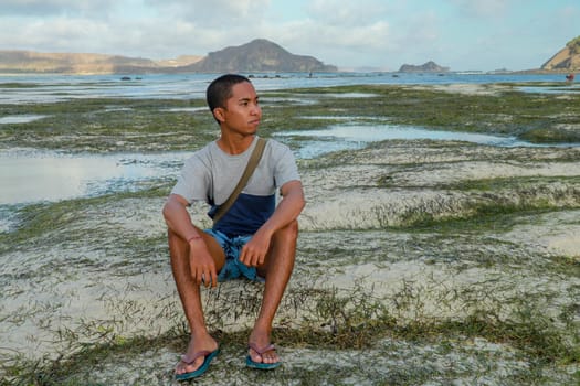 Relaxed young man meditating while sitting on the beach. Portrait of an attractive young man on a tropical beach. Teenager in a blue-gray T-shirt sitting on the beach.