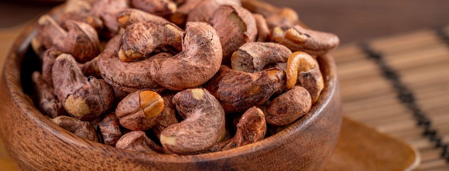 Cashew nuts with peel in a wooden bowl on wooden tray and table background, healthy raw food plate.