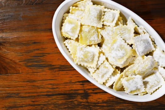 Heap of uncooked square ravioli in white bowl on red wooden table , ravioli with flour on surface