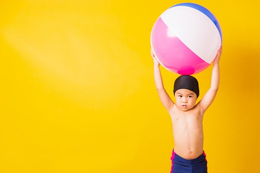 Summer vacation concept, Portrait Asian happy cute little child boy smiling in swimsuit hold beach ball, Kid having fun with inflatable ball in summer vacation, studio shot isolated yellow background