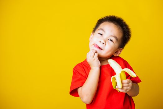 Happy portrait Asian child or kid cute little boy attractive smile wearing red t-shirt playing holds peeled banana for eating, studio shot isolated on yellow background