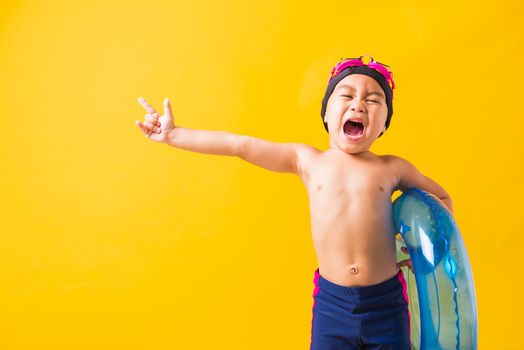 Summer vacation concept, Portrait Asian happy cute little child boy wear goggles and swimsuit hold blue inflatable ring, Kid hav fun point finger to side away, studio shot isolated yellow background