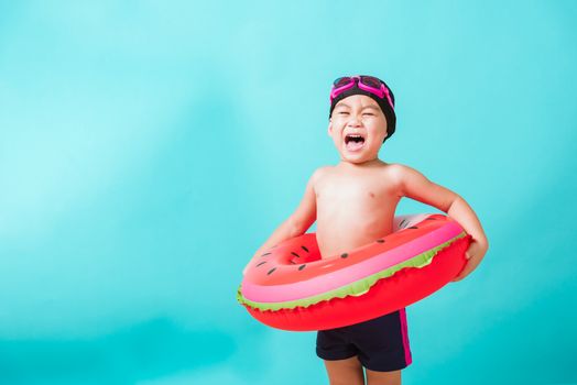 Summer vacation concept, Portrait Asian happy cute little child boy wear goggles and swimsuit hold watermelon inflatable ring, Kid having fun on summer vacation, studio shot isolated blue background