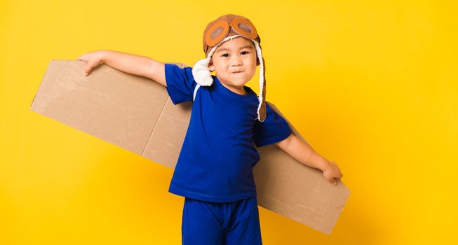 Happy Asian handsome funny child or kid little boy smile wear pilot hat playing and goggles with toy cardboard airplane wings flying, studio shot isolated yellow background, Startup freedom concept