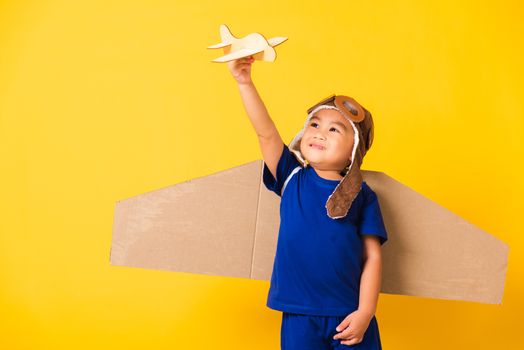 Happy Asian handsome funny child or kid little boy smile wear pilot hat play and goggles with toy cardboard airplane wings fly hold plane toy, studio shot isolated yellow background, Startup freedom