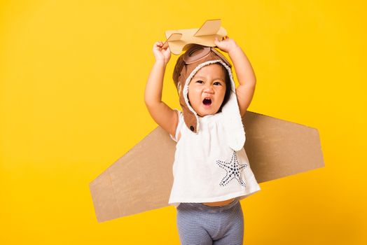 Happy Asian beautiful funny baby little girl smile wear pilot hat play and goggles with toy cardboard airplane wings fly hold plane toy, studio shot isolated yellow background, Startup freedom concept