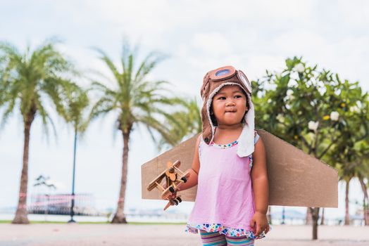 Happy Asian funny child or kid little girl smile wear pilot hat and goggles play toy cardboard airplane wing flying against summer sky cloud on trees garden background, Startup freedom concept