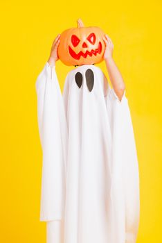 Funny Halloween Kid Concept, little cute child with white dressed costume halloween ghost scary he holding orange pumpkin ghost on hand, studio shot yellow on white background
