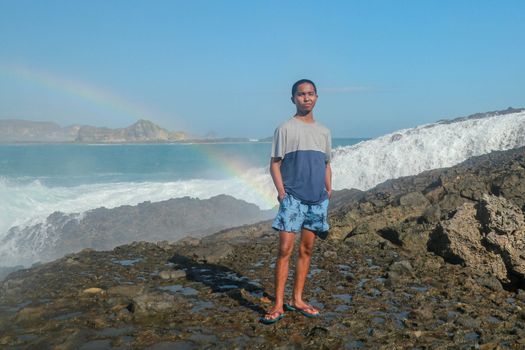 A man stands on a rocky shore and the waves crash against a cliff. Rainbow phenomenon in water fog. Waves hitting round rocks and splashing.