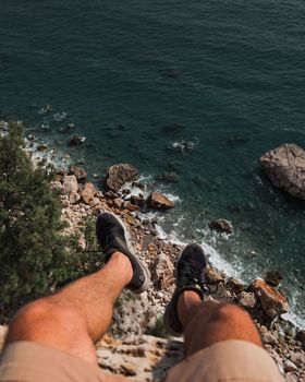 Legs of man are hanging from edge of cliff over the sea and rocks.