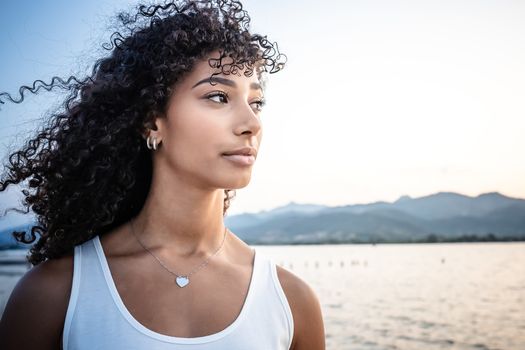 Beautiful Hispanic young woman head and shoulders portrait with long curly dark hair in white undershirt looks to the sun at dusk with lake and mountains background - Human beauty and that of nature