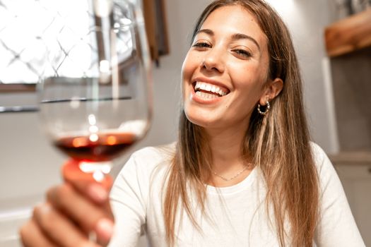 Cute alone long hair blond caucasian smiling woman toasting with red wine glass at home looking at the camera - New normal distancing technology social relations for Coronavirus