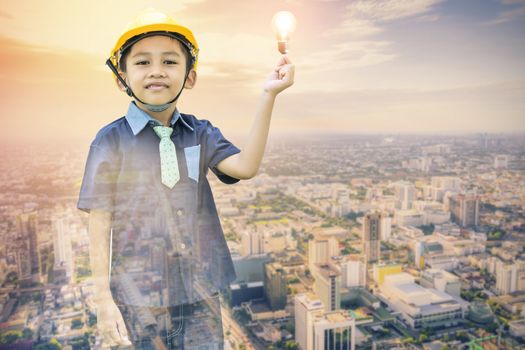 Double exposure of the Asian boy dressed as an engineer wearing a safety hat holding a light bulb on a cityscape backdrop-Imagination and creativity concepts.