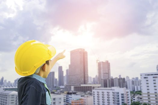 A boy dressed as an engineer wearing a safety hat pointing his hand on the cityscape with sunset background-concepts for the future is growing up to be an engineer.