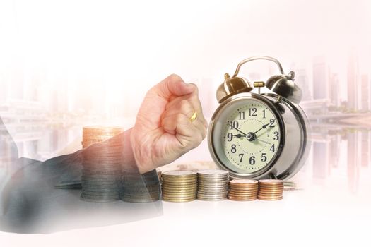 Double exposure of hand of business fist with a row of stack money coins and analog clock on the blurred cityscape background concept for business finances and saving money.