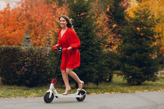 Young woman with electric scooter in red dress at the autumn city park.
