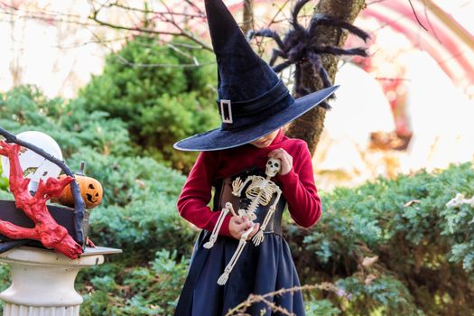 Little girl in a witch costume holding a skeleton on a halloween party in the garden