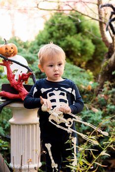 Little boy in a skeleton costume holding a skeleton on a halloween party in the garden