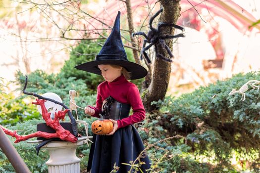 Little girl in a witch costume holding a skeleton on a halloween party in the garden