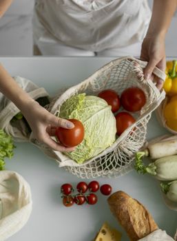 Woman came back from a market and unpacks a reusable grocery bag full of vegetables on a kitchen at home. Zero waste and plastic free concept. Girl is holding mesh cotton shopper with vegetables