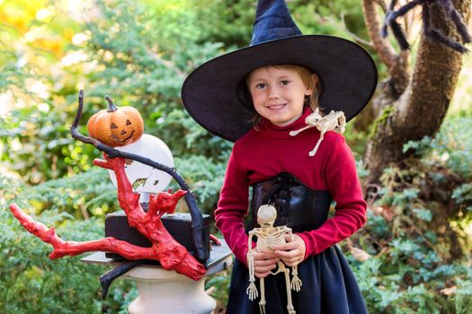 Little girl in a witch costume holding a skeleton on a halloween party in the garden