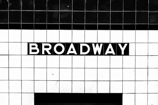 Black and white image of Broadway subway stop sign made of tiles opposite the platform