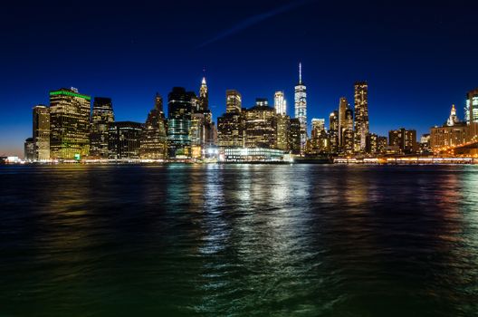 View of illuminated Manhattan skyline at twilight, with skyscraper lights reflected on the river. New York City, USA