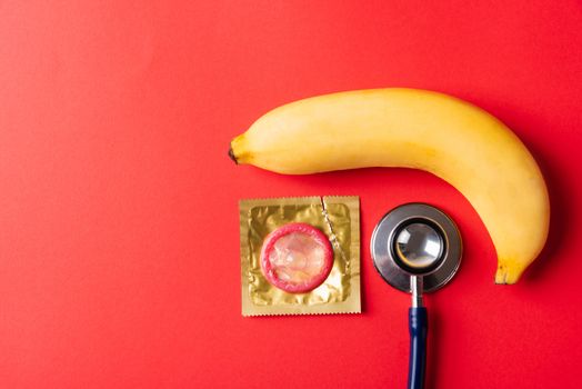 World sexual health or Aids day, Top view flat lay condom in wrapper pack, banana and doctor stethoscope, studio shot isolated on a red background, Safe sex and reproductive health concept