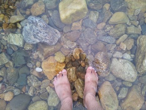 Top view of transparent water surface with human feet visible in it. Copy space for background and texture. Transparent water texture with view of stones under water.