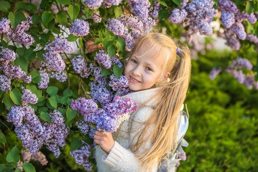 Little blonde girl smelling lilac flowers in beautiful sunny day. Cute girl outdoors in the blooming garden. Kid in nature in summer