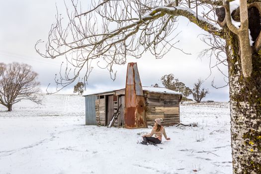 Woman relaxing in snowy field with old rustic barn made of recycled timber planks, pieces of corrugated iron and rusting metal sheets